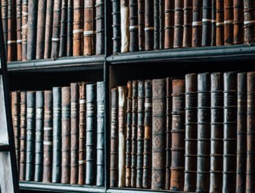 A tall wooden bookshelf filled with old, worn books separated by thin white horizontal labels stands in the study of a prominent lawyer, with a wooden ladder leaning against it.