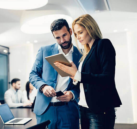 A man in a blue suit and a woman in a black blazer are discussing information on a tablet in an office setting.