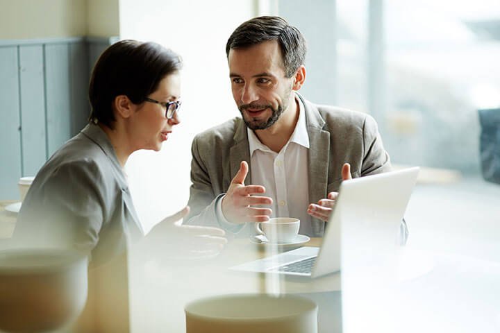 Two professionals in business attire are having a conversation at a café table, with a laptop and coffee cups in front of them; one of them appears to be an employer seeking legal advice from the other, possibly a lawyer.