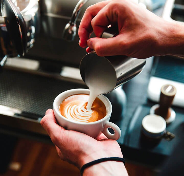 A barista, under the watchful eye of their employer, pours steamed milk into a cup of espresso, creating intricate latte art.