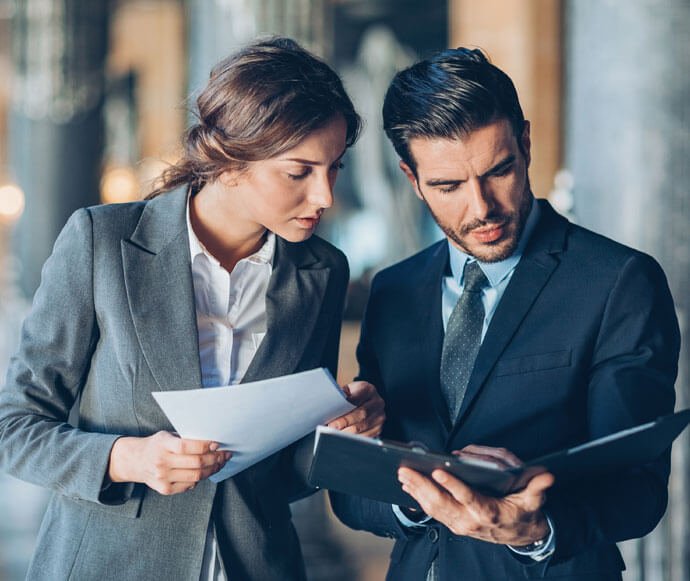 Two business professionals, a woman and a man, are standing and looking over important documents together. The employer is holding a tablet while the lawyer appears to be discussing significant details with him.