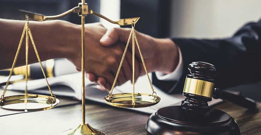 Two people shaking hands over a contract near a gavel and scales of justice, symbolizing a legal agreement or settlement.