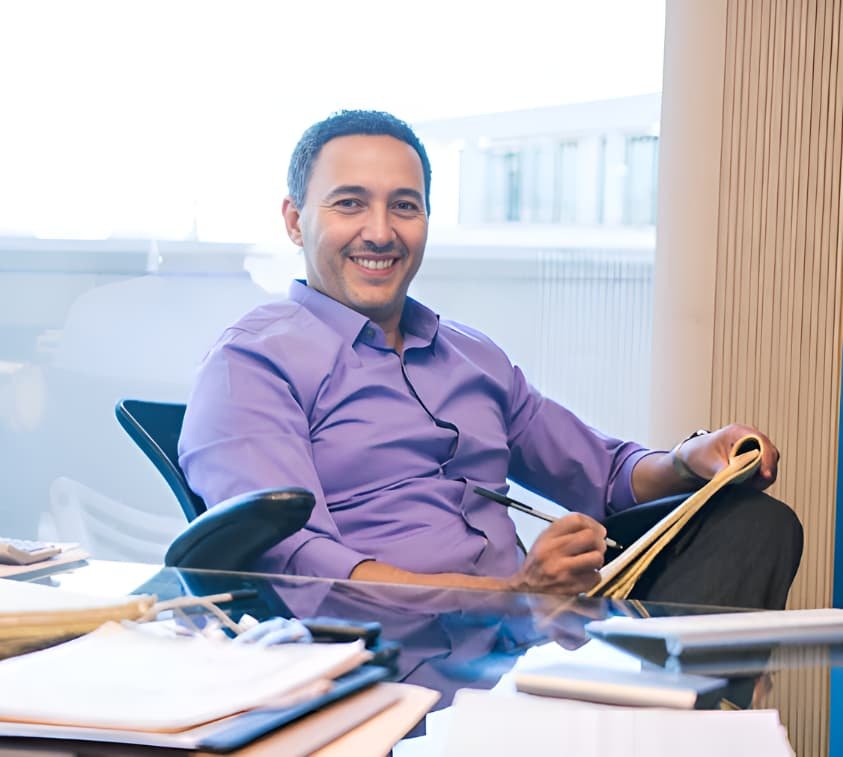 A man in a purple shirt smiles while sitting at a desk, holding a notebook and pen.