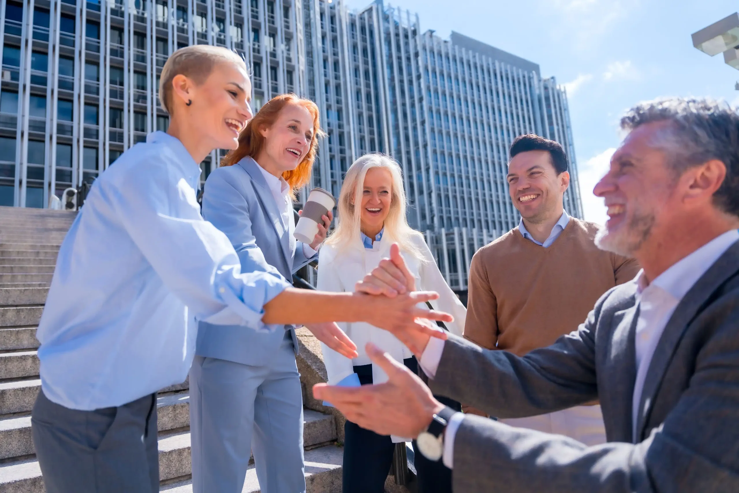 A group of five people, dressed in business attire, stand outdoors near a modern glass building. They are smiling and gesturing amicably, with some individuals shaking hands.