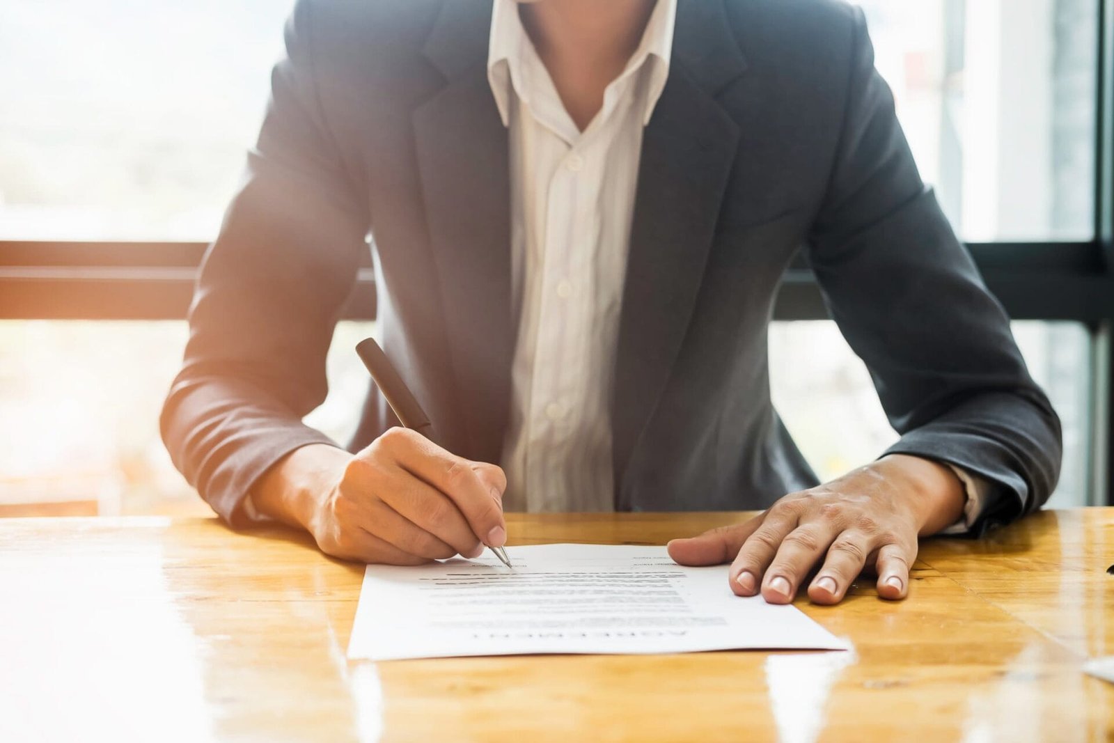 Person in a blazer signing a document on a wooden desk, with natural light coming through the window in the background, possibly seeking guidance from an employment lawyer offering free consultations.