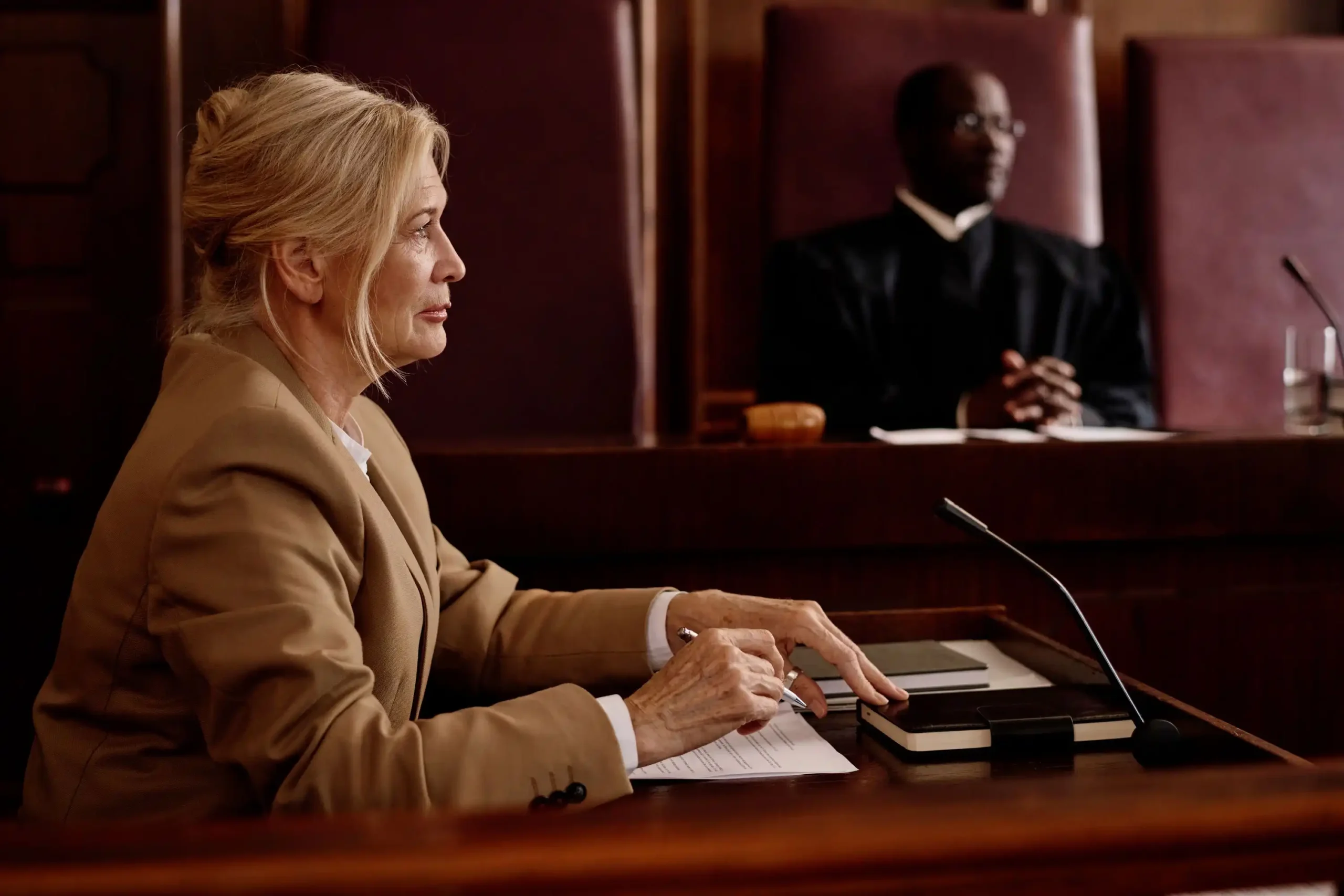 A woman in a tan blazer sits at a desk in a courtroom, with a judge in a black robe seated behind her.