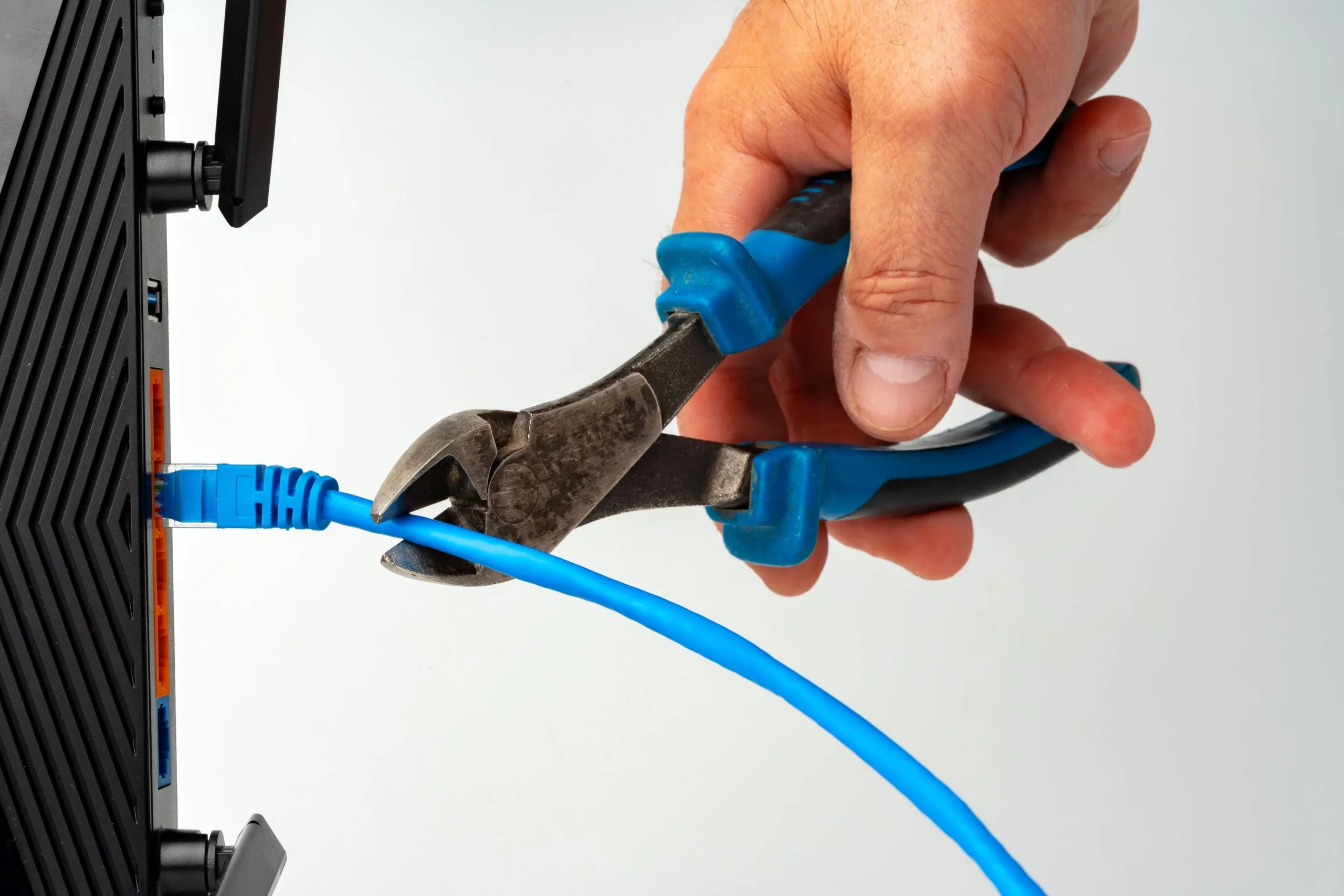 A hand holding blue-handled wire cutters prepares to cut a blue Ethernet cable connected to a black device.