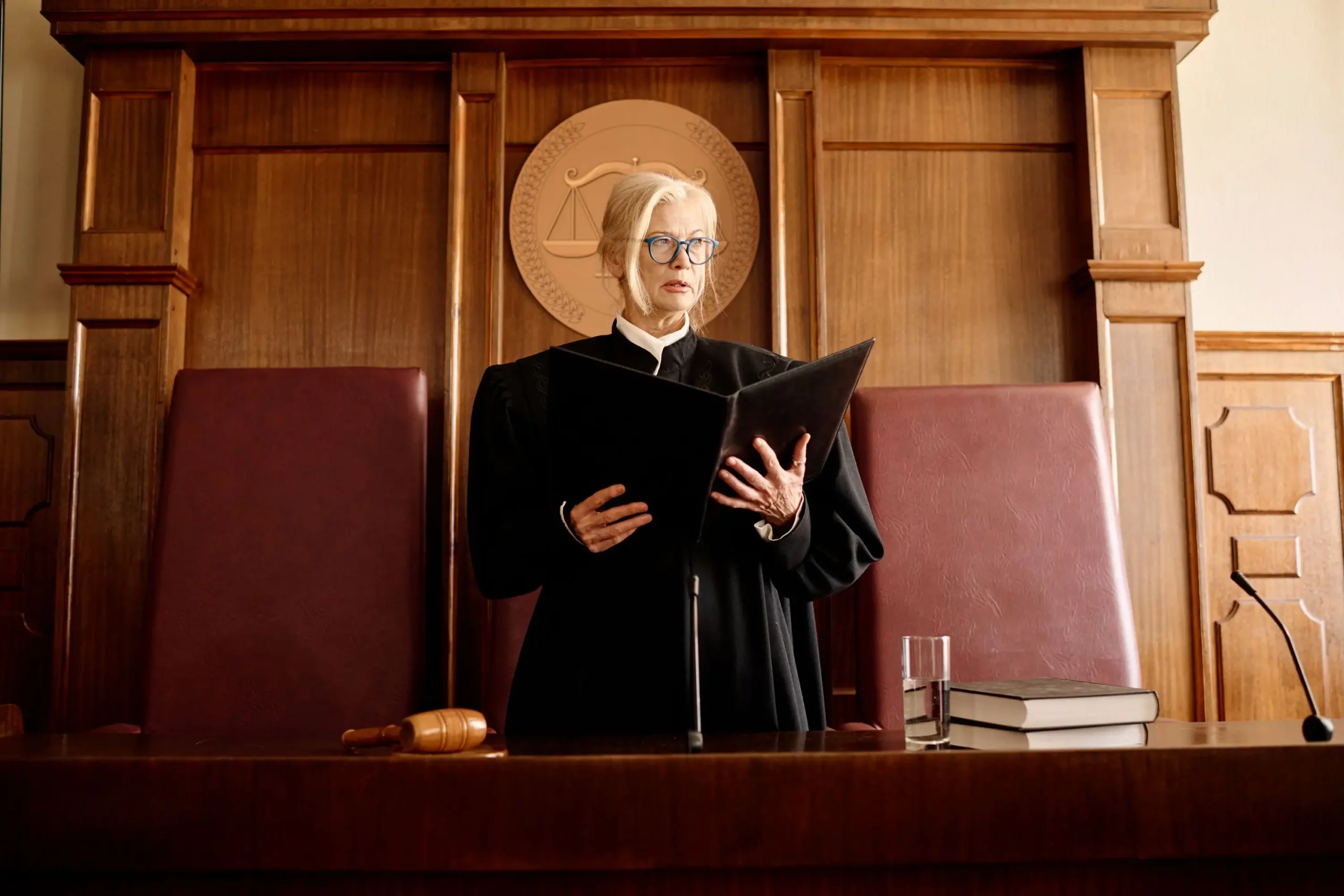 A person wearing glasses and a judge's robe stands at a courtroom bench, holding a black folder. A gavel, a glass of water, and a book are on the desk in front.