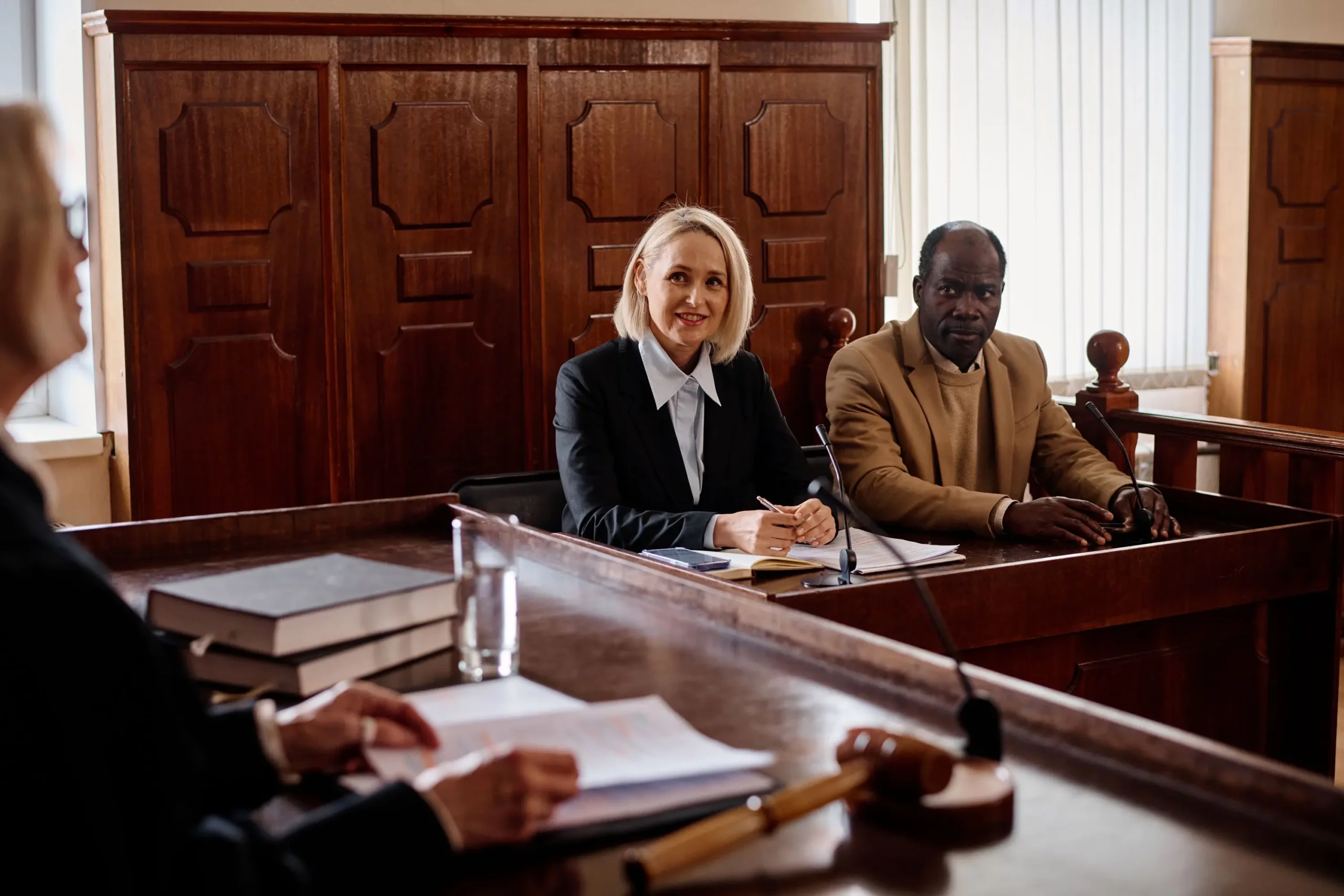 Two people, one woman in a suit and one man in a jacket, sit at a courtroom table. A judge's gavel and documents are in the foreground.