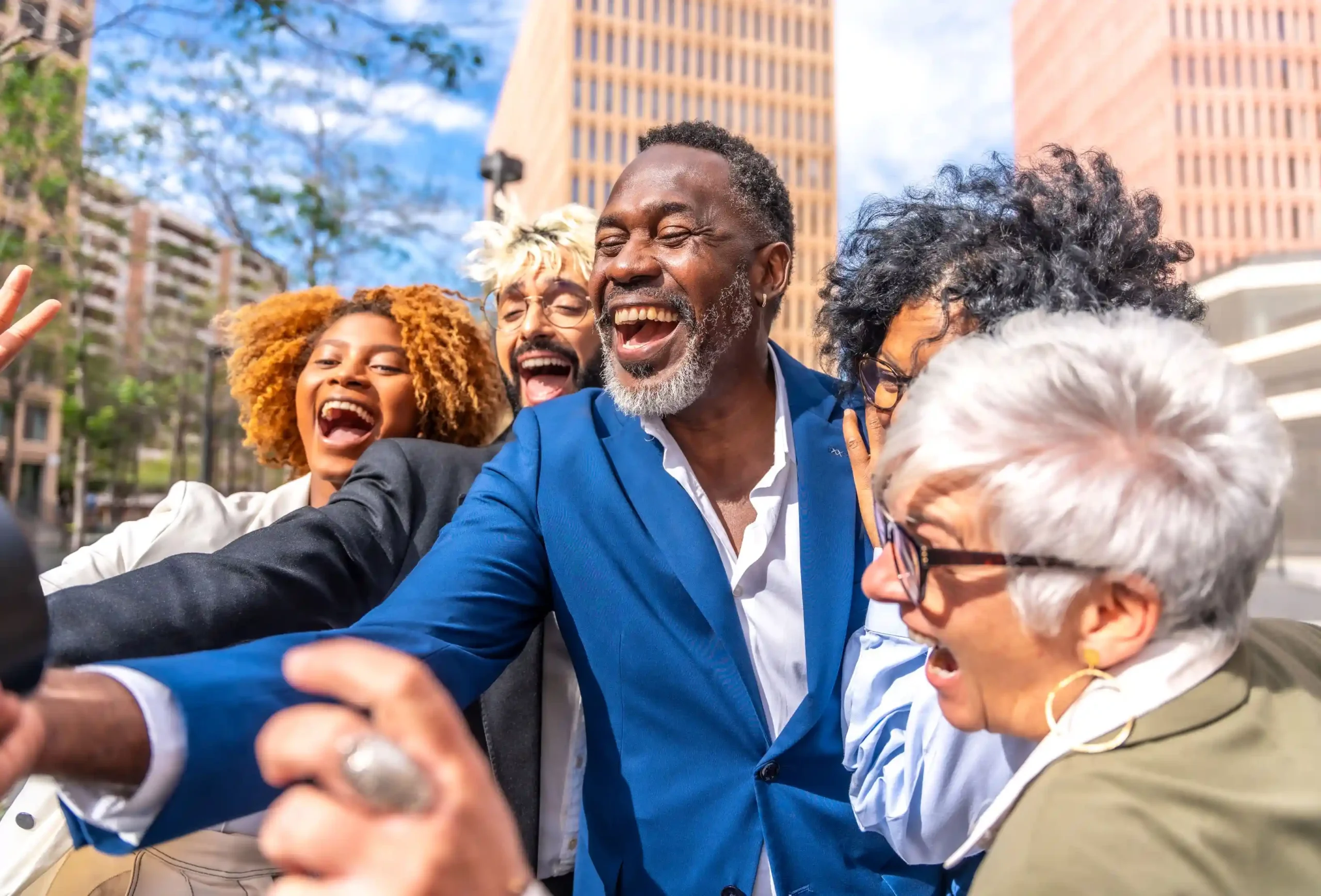 A group of diverse, smiling adults wearing business attire take a selfie together in an urban environment with tall buildings in the background.