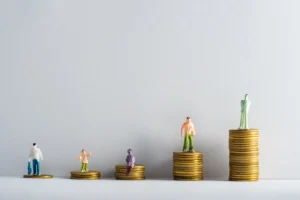 Four miniature figures stand on stacks of coins of varying heights, representing differing levels of wealth or financial status.