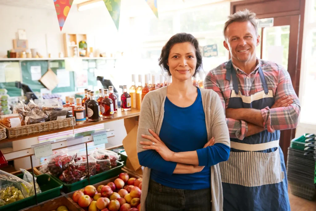 Two people stand smiling inside a grocery store with fruits, vegetables, and various products on shelves behind them. One is wearing an apron.