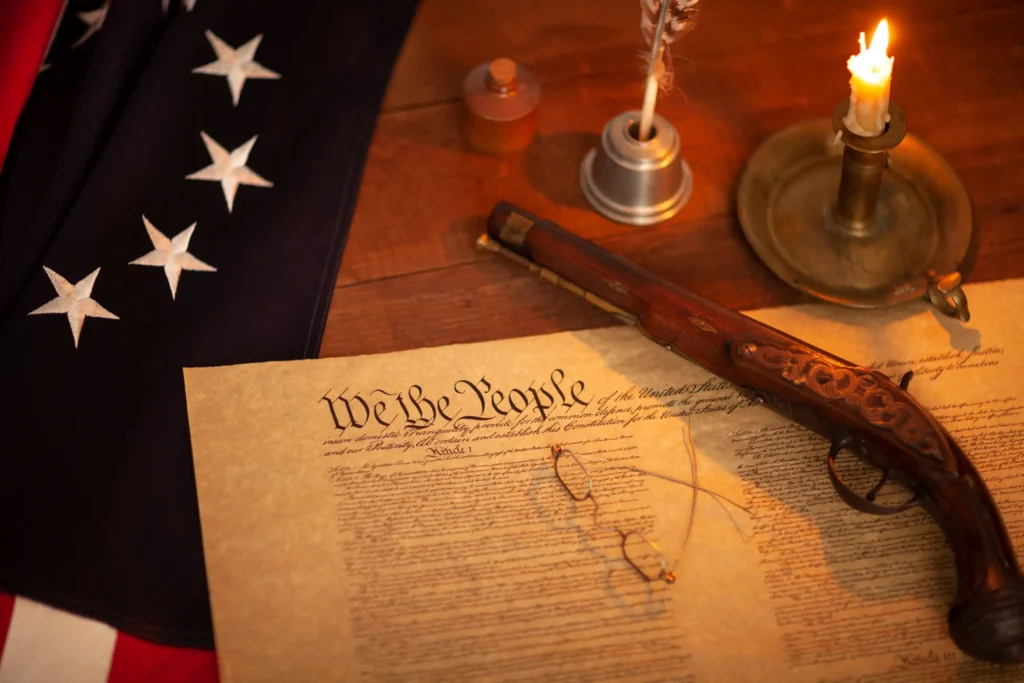 A historical document with the words "We the People," a quill, spectacles, a candle, a pistol, and part of a flag with stars are placed on a wooden table.