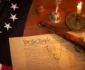 A historical document with the words "We the People," a quill, spectacles, a candle, a pistol, and part of a flag with stars are placed on a wooden table.