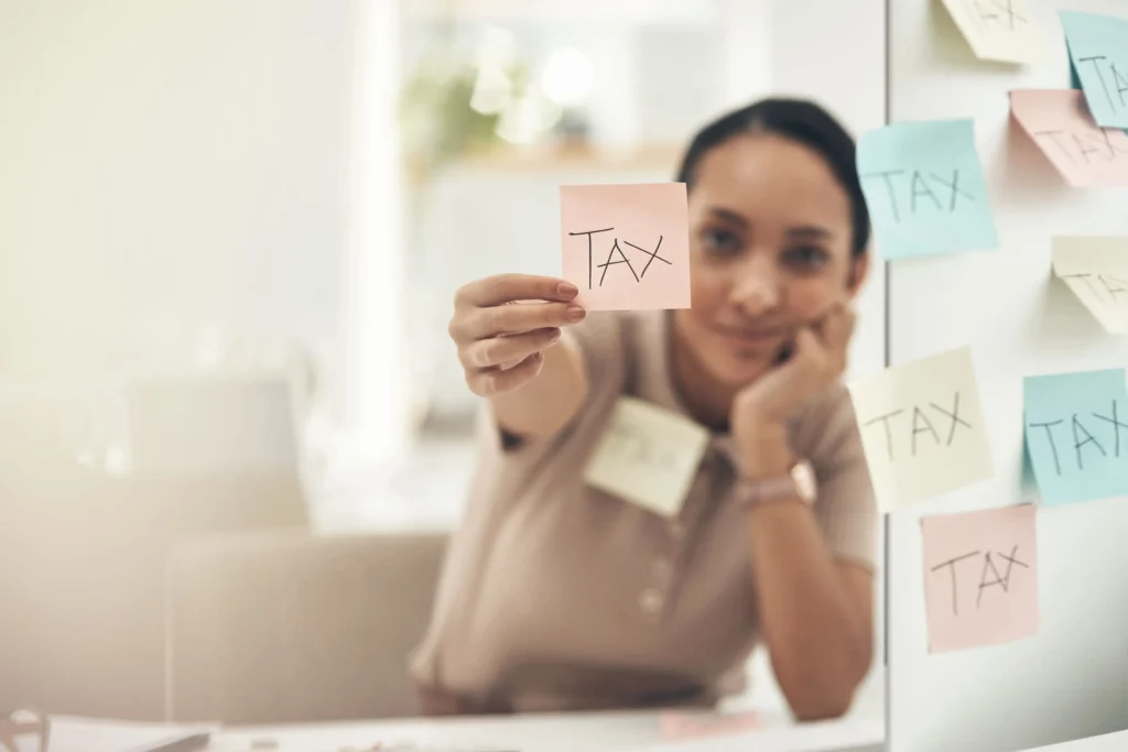 A woman holds a pink sticky note with the word "TAX" written on it, facing the camera. She is surrounded by other sticky notes with the same word, attached to a wall beside her.