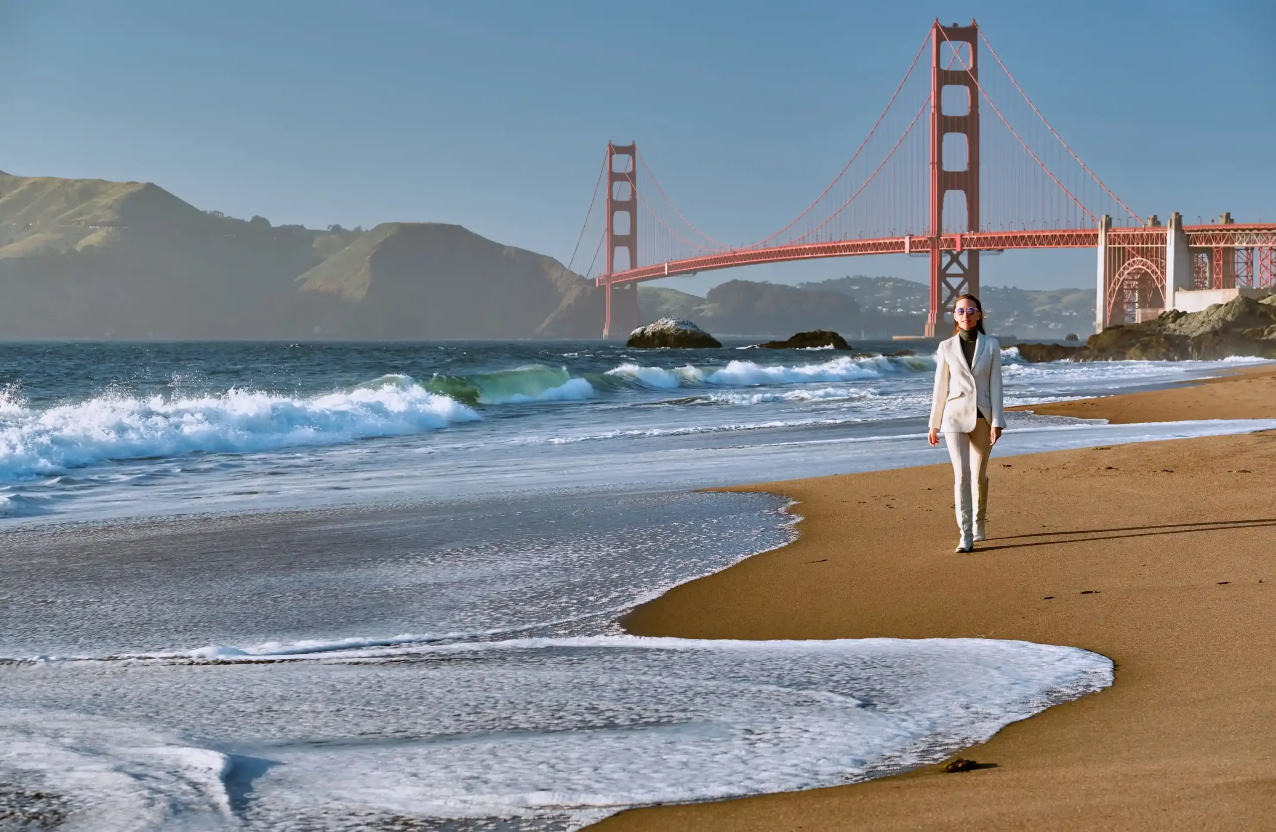 A person walks along the sandy beach, with waves rolling in and the Golden Gate Bridge in the background under a clear sky.