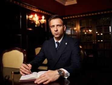 A man in a suit sits at a wooden table with an open book in a dimly lit room with bookshelves in the background.