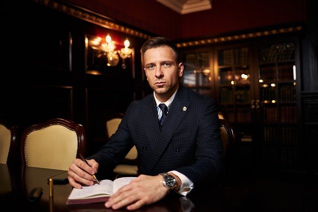 A man in a suit sits at a wooden table with an open book in a dimly lit room with bookshelves in the background.